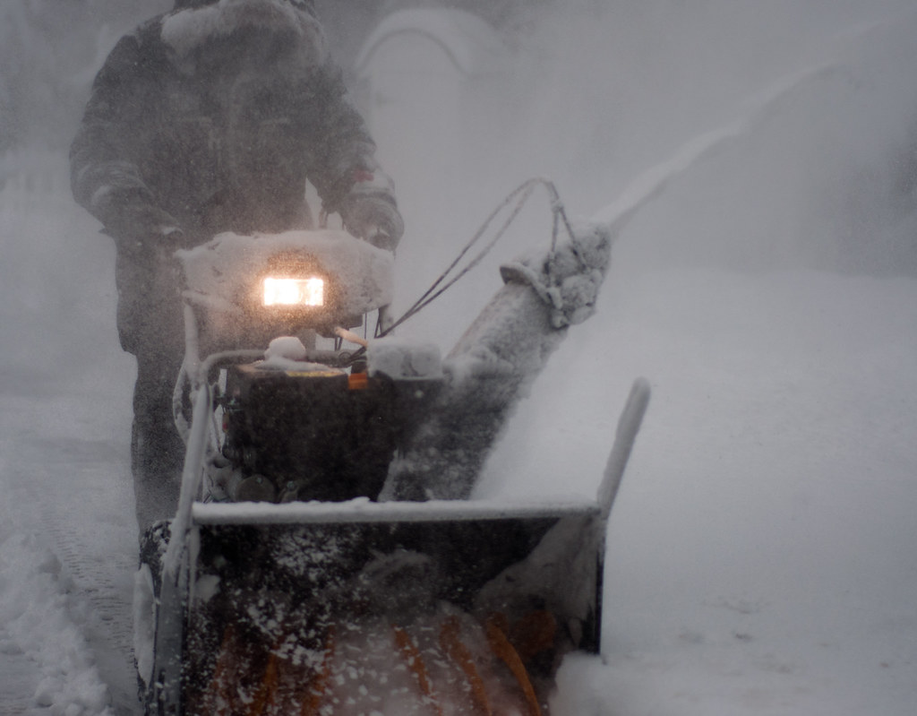 A person pushing a walk-behind two-stage snow blower through heavy blowing snow, its headlight on and snow arcing from the chute